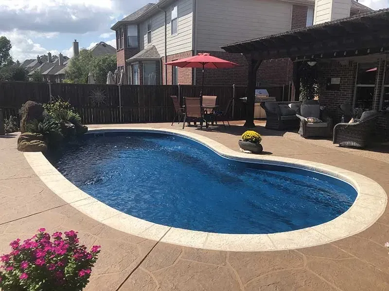 Backyard pool with blue water, tan deck, red umbrella, and patio seating beside a house
