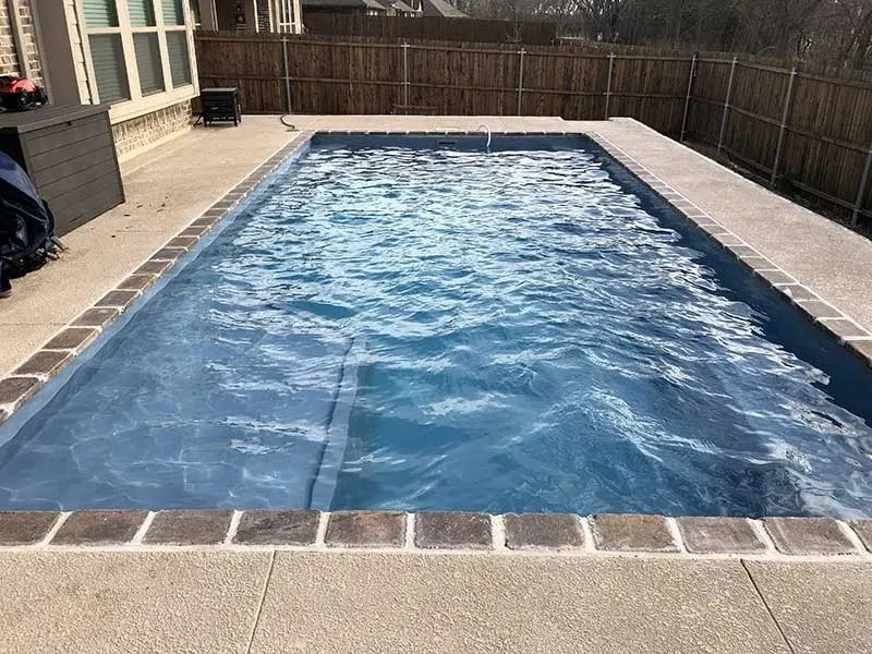 Rectangular backyard swimming pool with blue water, surrounded by a paved deck and wooden fence