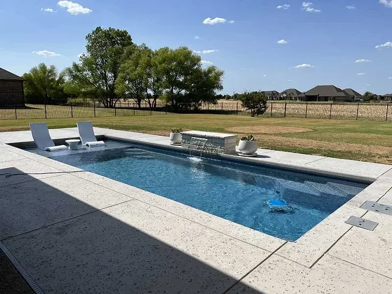 Backyard patio with a rectangular in-ground pool, lounge chairs, and a grassy yard under a blue sky
