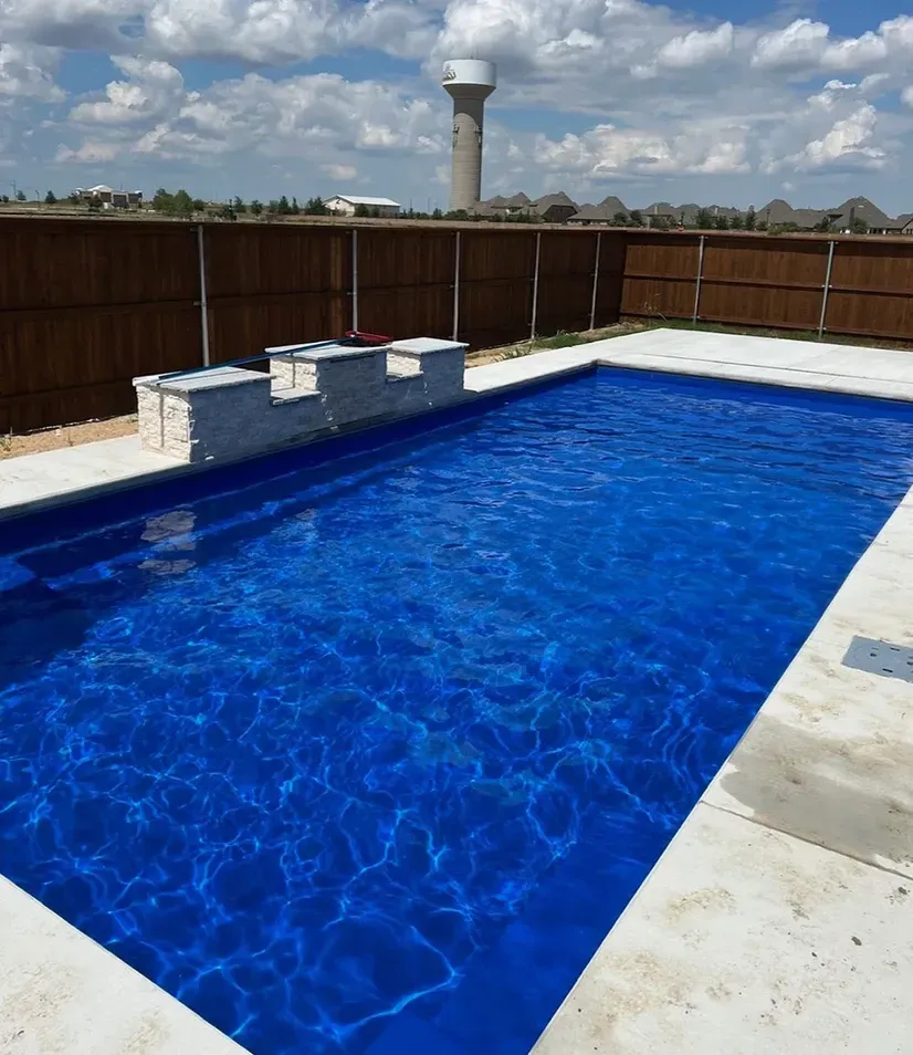 Backyard swimming pool with blue water, white deck, and brown fence under a cloudy sky