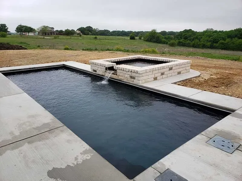 Rectangular black-lined pool beside a raised hot tub on a concrete patio, with grassy fields beyond