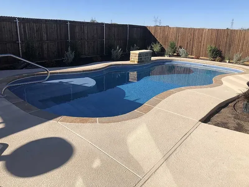 Backyard swimming pool with curved concrete deck, blue water, and wooden privacy fence