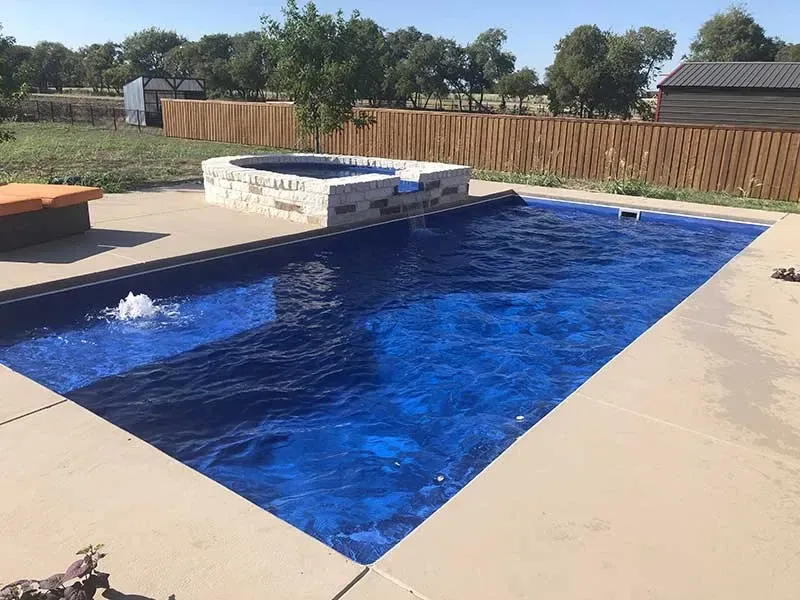 Rectangular backyard pool with blue water, stone deck, and raised spa beside a wooden fence.