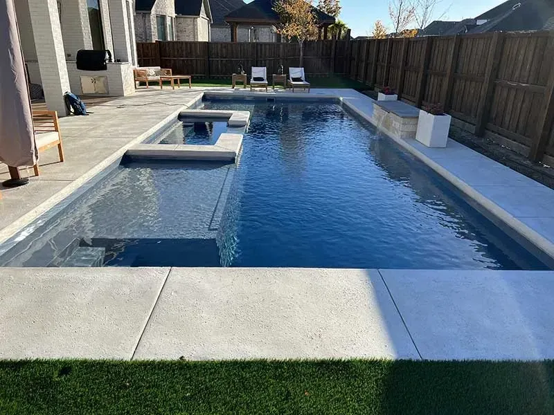 Backyard swimming pool with a shallow spa ledge, concrete deck, and backyard fencing in sunlight