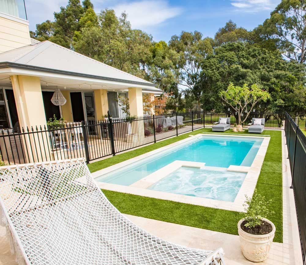 Backyard patio with a rectangular swimming pool, white house, and lounge chairs under trees.