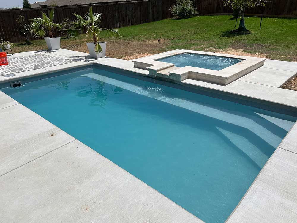 Backyard with a rectangular blue swimming pool and a raised square spa on a concrete patio.