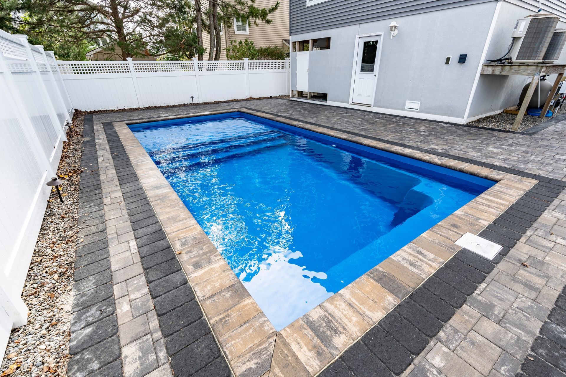Rectangular in-ground pool with blue water, bordered by brick pavers and a white fence in a backyard