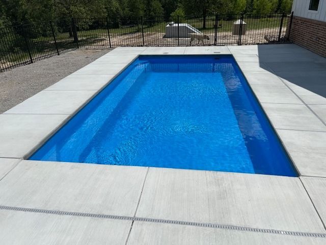 Outdoor rectangular swimming pool with blue water and light gray deck, bordered by a black fence and trees