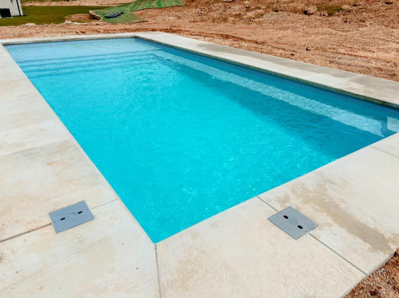 Rectangular outdoor swimming pool with bright blue water, set in a concrete deck and dirt landscaping