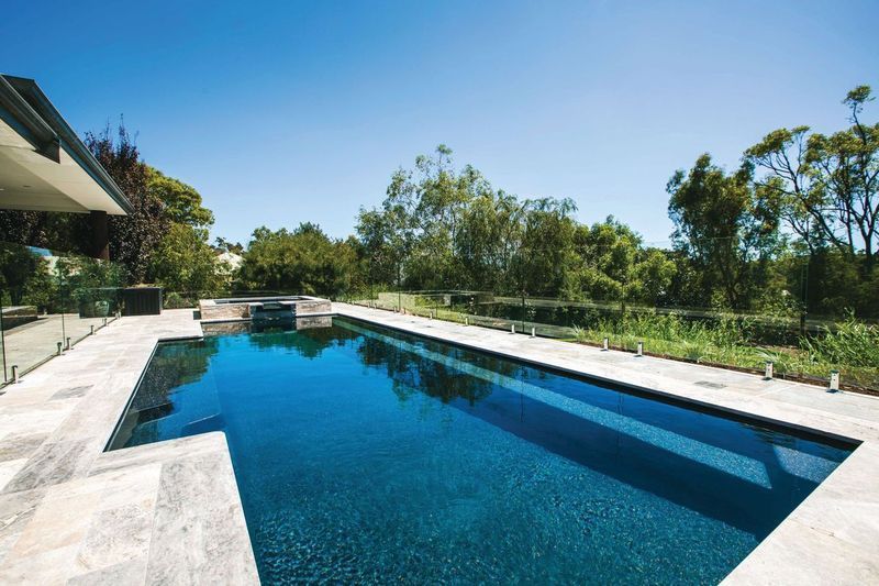 Rectangular outdoor swimming pool beside a modern house, surrounded by trees under a clear blue sky