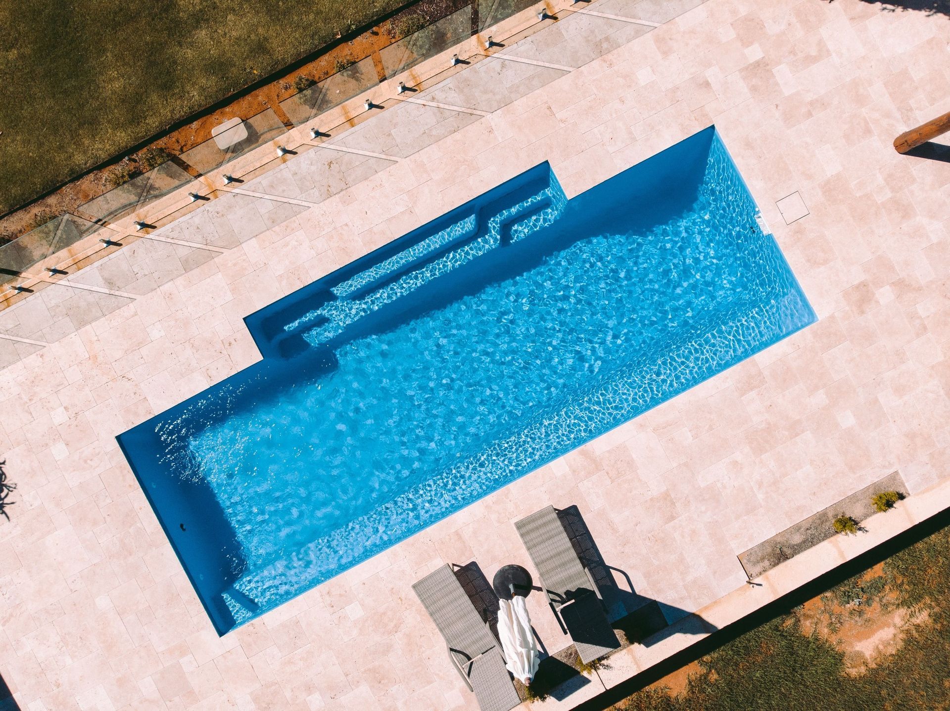 Aerial view of a rectangular blue swimming pool beside a patio with lounge chairs and surrounding grass.