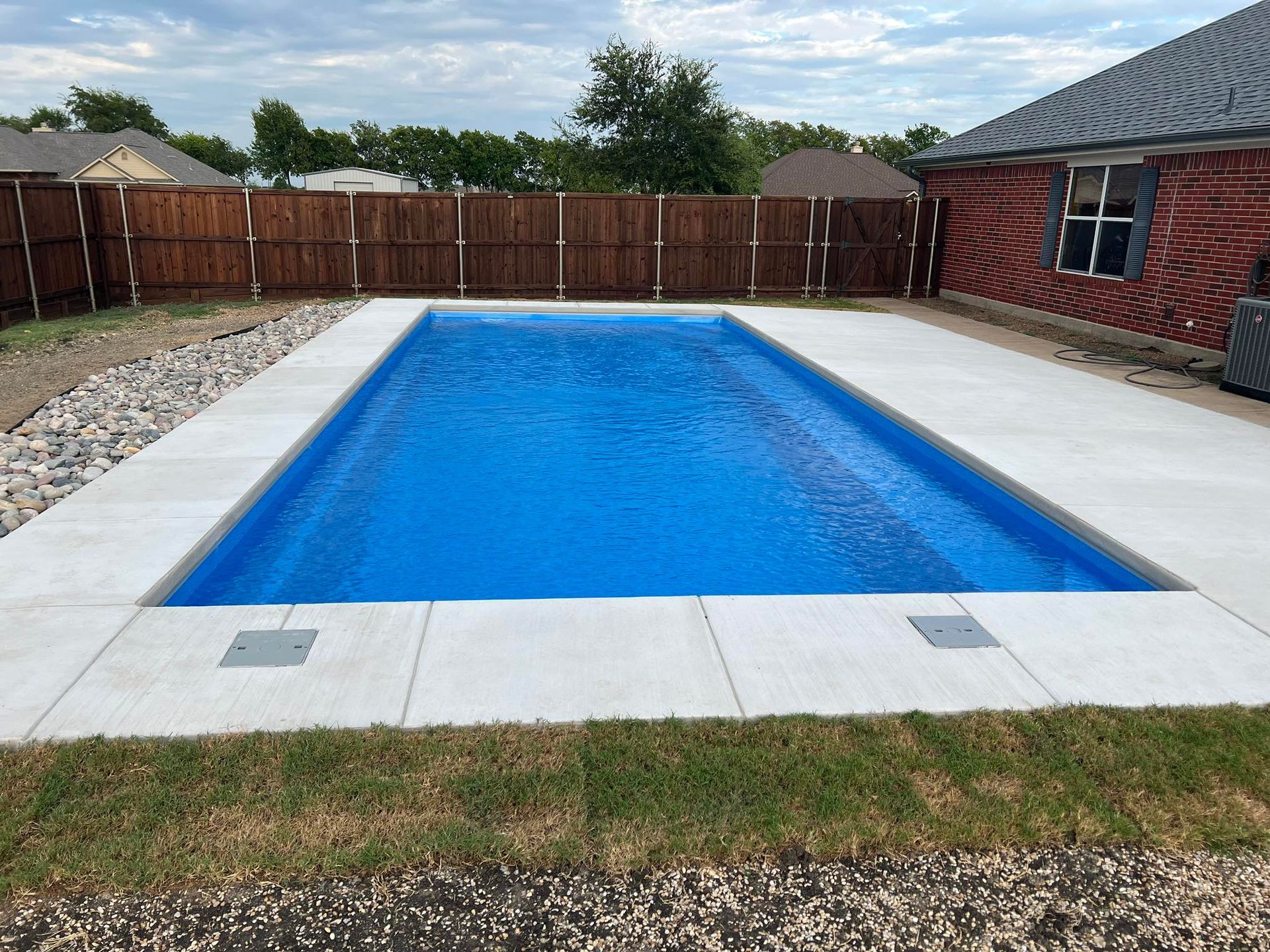 Rectangular blue swimming pool in a fenced backyard with stone coping and grass lawn