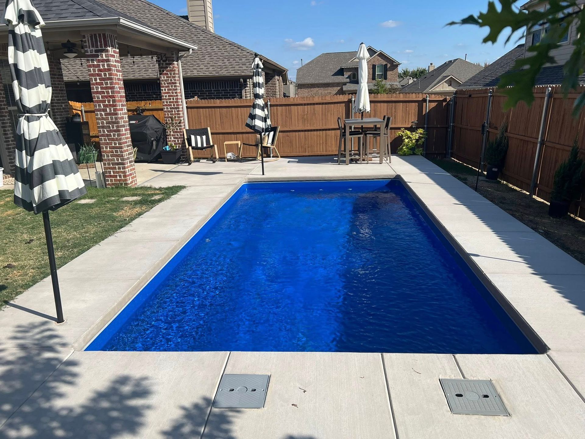 Backyard swimming pool with blue water, brick patio, and fence under a sunny sky