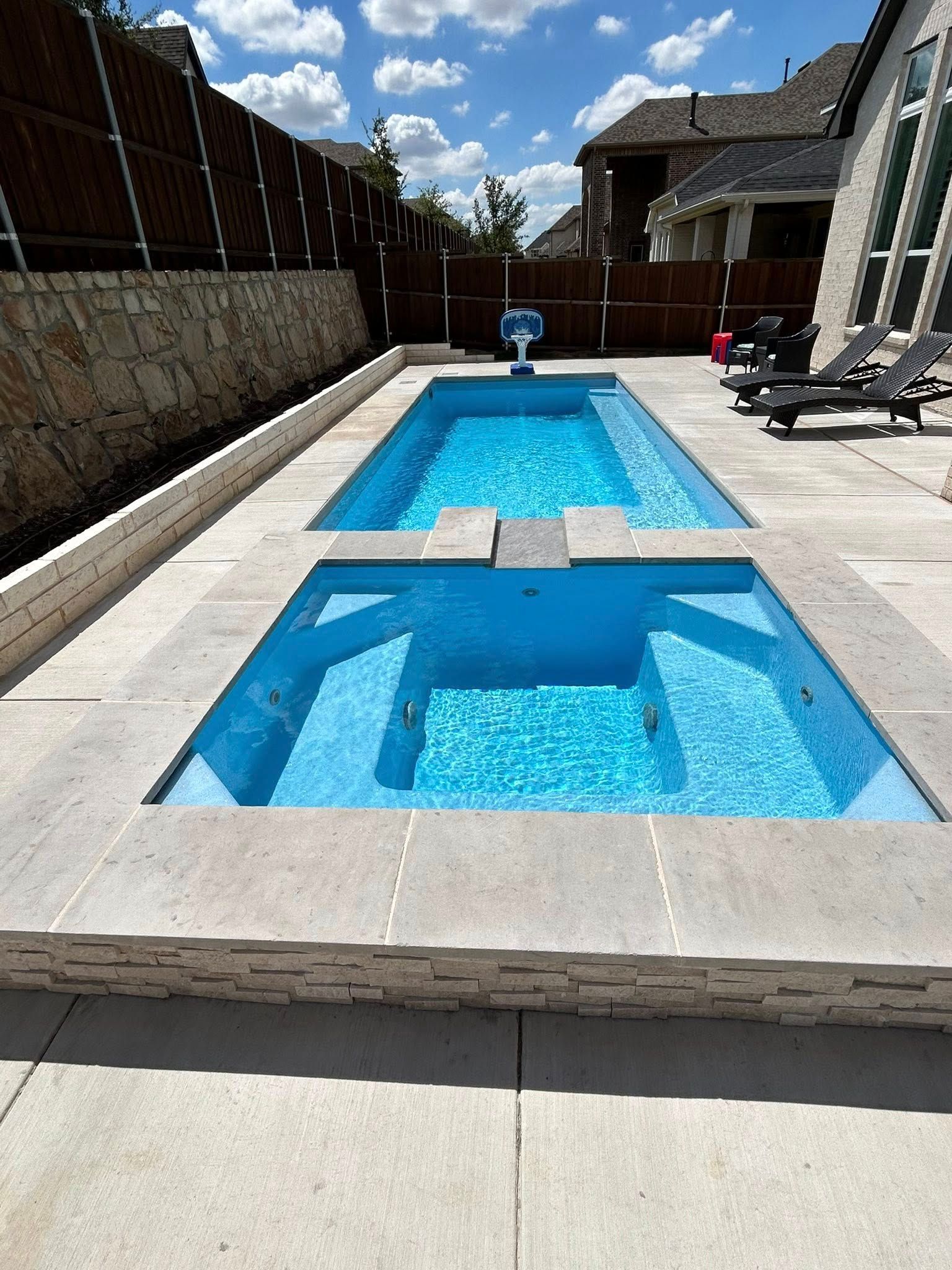 Rectangular backyard pool with blue water, concrete deck, lounge chairs, and a partly cloudy sky