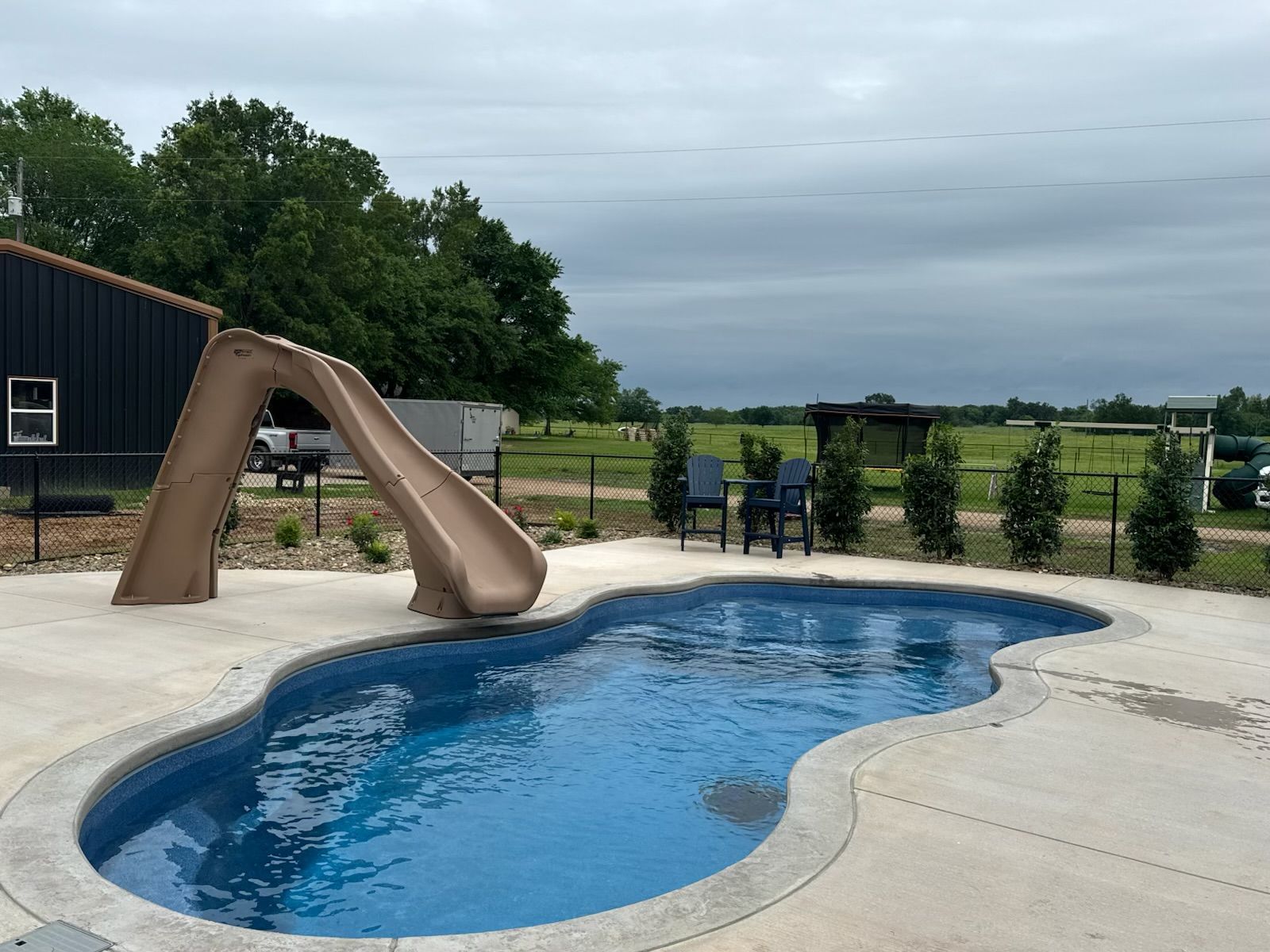 Backyard pool with tan slide, concrete deck, and fenced green landscape under cloudy sky