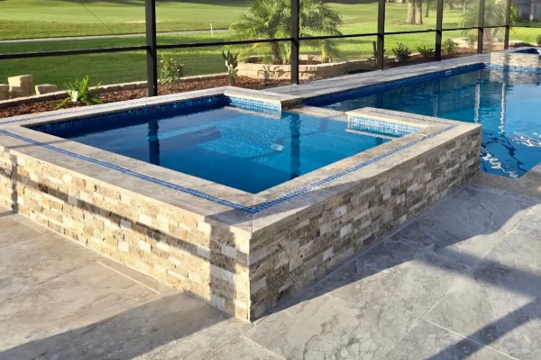 Stone-walled outdoor hot tub beside a blue swimming pool on a sunny patio