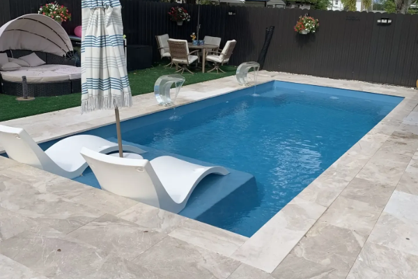 Rectangular backyard pool with white lounge chairs, stone patio, and black privacy fence