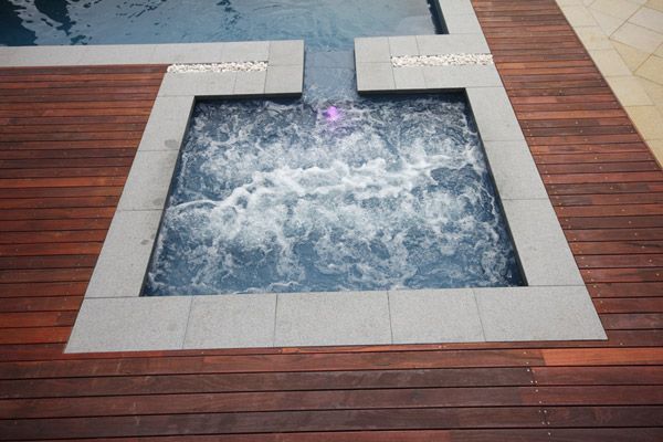 Top-down view of a bubbling square hot tub inset in a wooden deck, with blue water and white stone edging
