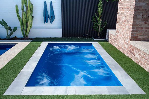 Modern backyard with a rectangular blue swimming pool, white border, and brick wall beside a patio.