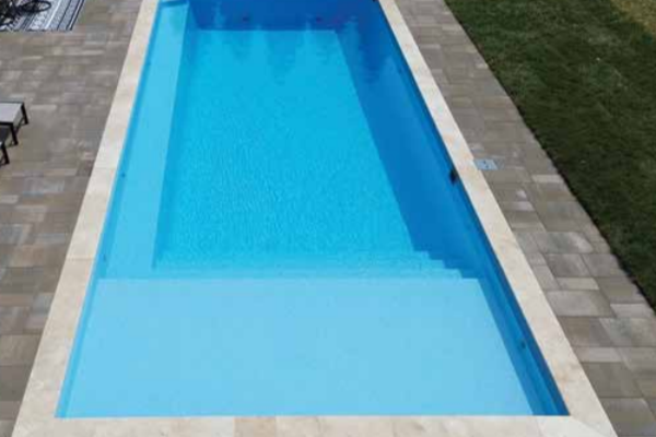 Long rectangular blue swimming pool bordered by light stone tiles and grass