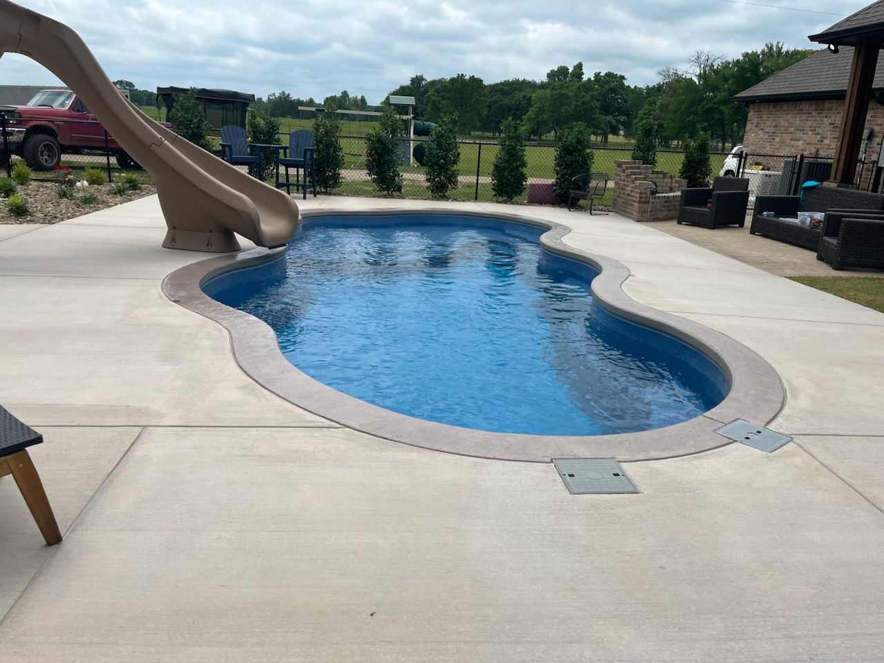 Backyard swimming pool with a tan slide and surrounding patio, bordered by landscaped shrubs.