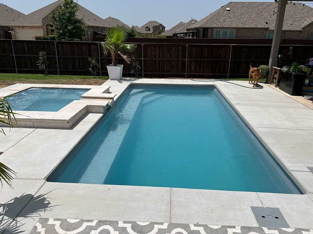 Backyard swimming pool with blue water and a raised spa, surrounded by white stone patio and fence
