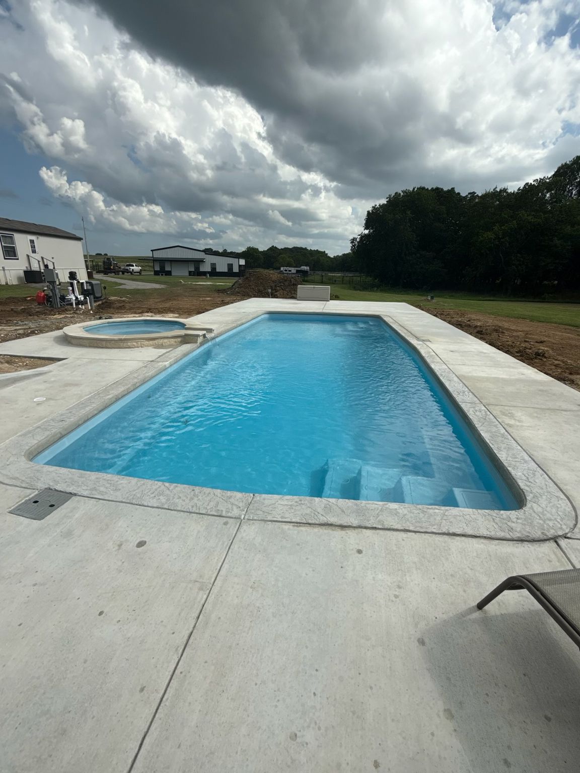 Outdoor swimming pool with blue water under a cloudy sky, surrounded by a concrete deck and grassy yard
