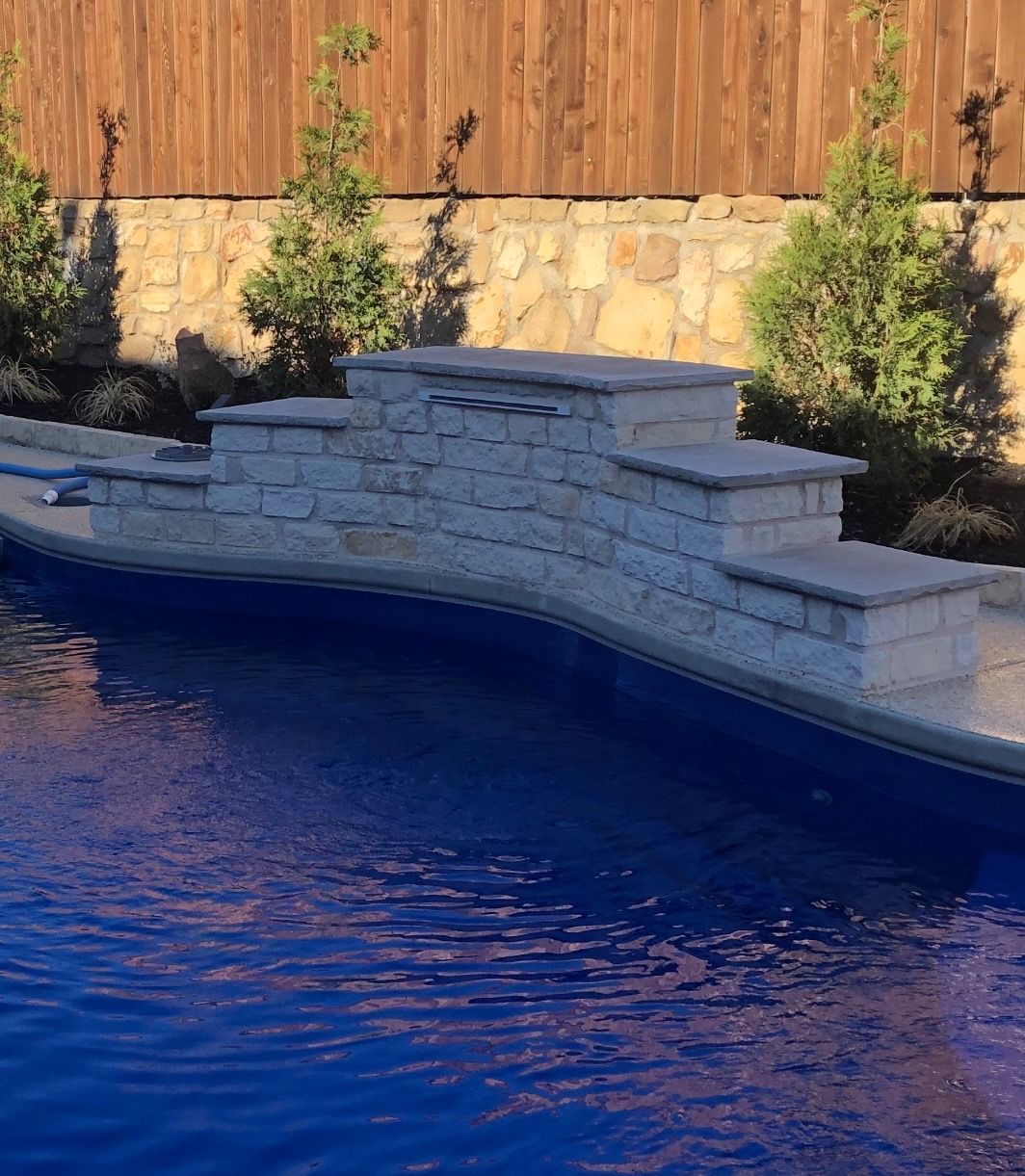 Backyard pool with a stone waterfall and steps against a wooden fence and shrubs