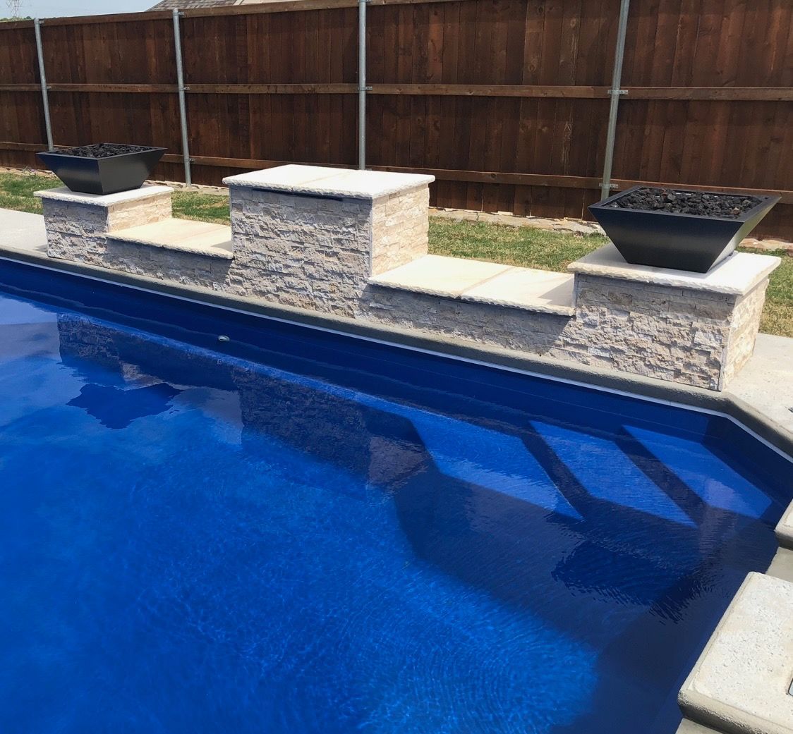 Backyard swimming pool with blue water, stone coping, and a wooden privacy fence.