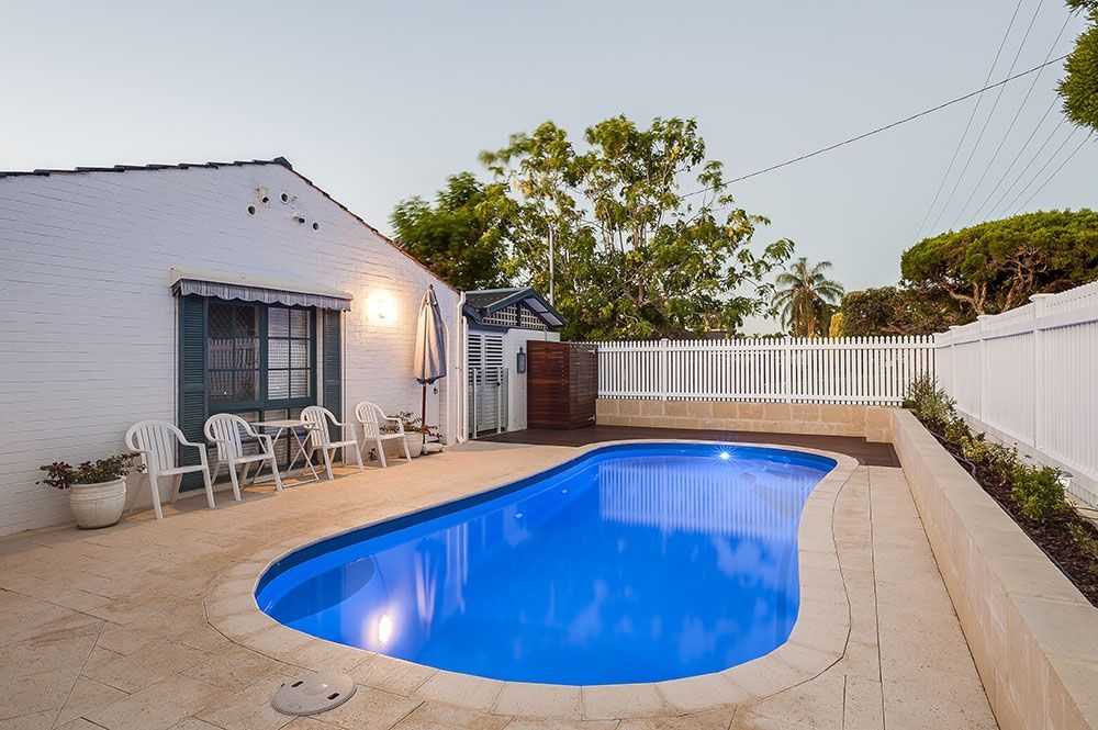 Backyard patio with a lit swimming pool, white fence, and house seating area at dusk