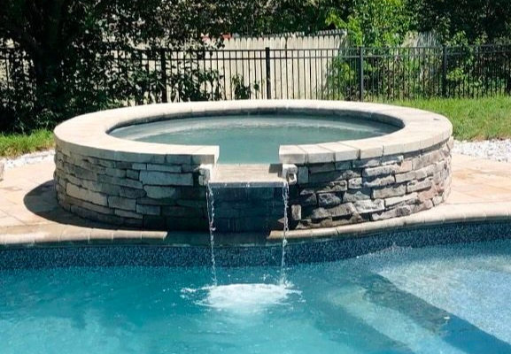 Round stone hot tub spilling water into a blue swimming pool outdoors
