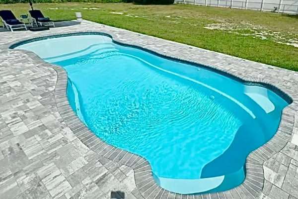 Curved backyard swimming pool with blue water and gray stone patio, bordered by grass and lounge chairs