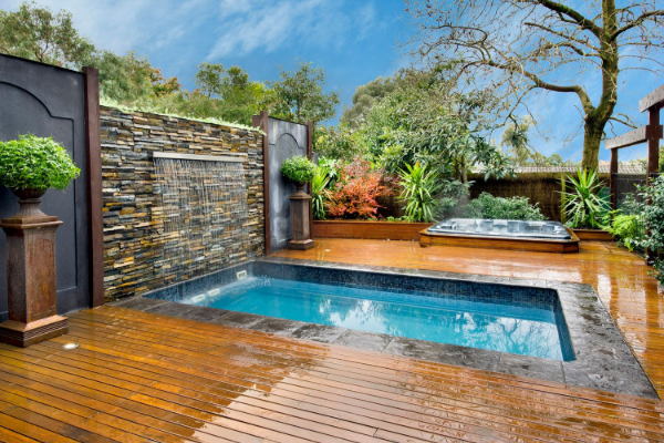 Modern backyard pool with wooden deck, stone wall, hot tub, and lush landscaping under a blue sky