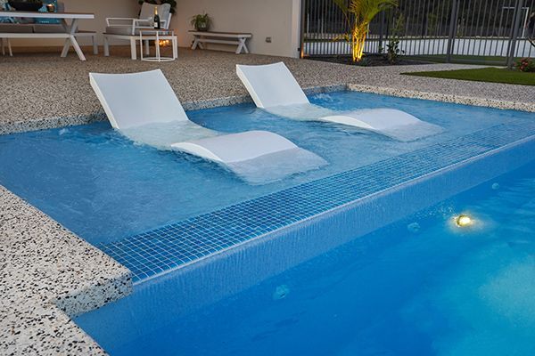 Poolside lounge chairs in a shallow resort pool at night with blue lighting