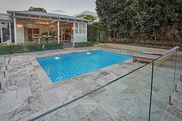 Modern backyard with a blue pool, stone patio, glass railing, and a house in the background