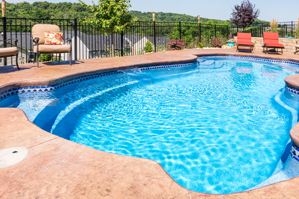 Curved backyard swimming pool with blue water, tan stone deck, lounge chairs, and a metal fence behind it