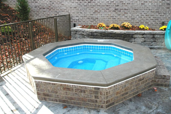 Small octagonal above-ground hot tub with blue water on a brick patio by a fence and brick wall