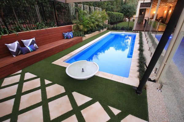 Modern backyard with a narrow blue pool, tiled patio, and wooden bench seating with cushions