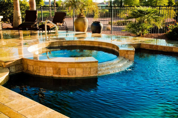 Circular stone hot tub beside a blue pool with a waterfall in a landscaped backyard