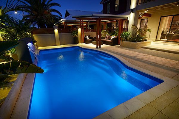Illuminated backyard swimming pool at night with patio seating and tropical plants