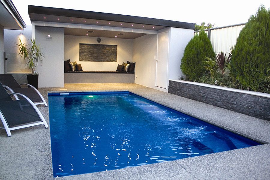 Modern backyard pool with blue water, lounge chair, and covered patio seating area at dusk
