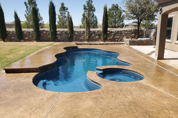 Backyard swimming pool with curved blue water and tan concrete patio, bordered by trees and stone wall