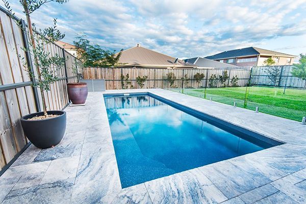 Modern backyard with a rectangular blue pool, stone patio, and fenced lawn under a partly cloudy sky