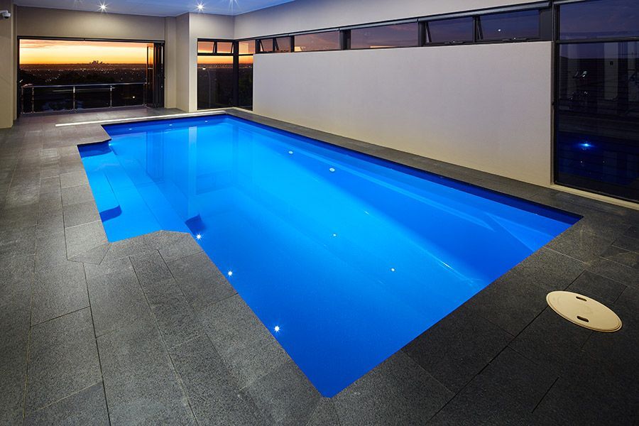 Indoor rooftop pool with bright blue water, dark tile deck, and sunset view through large windows