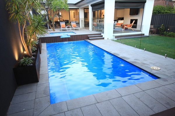 Modern backyard with a rectangular blue swimming pool, stone patio, and lit outdoor lounge area
