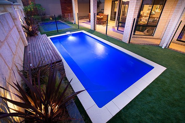 Modern backyard patio with a rectangular blue-lit pool, wooden bench, and house exterior at dusk