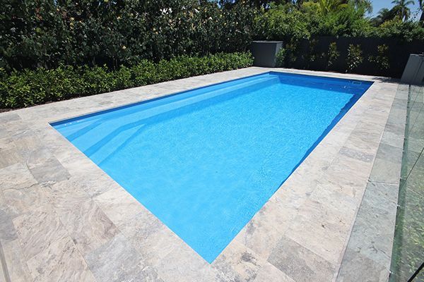 Rectangular outdoor swimming pool with bright blue water and stone decking, surrounded by greenery