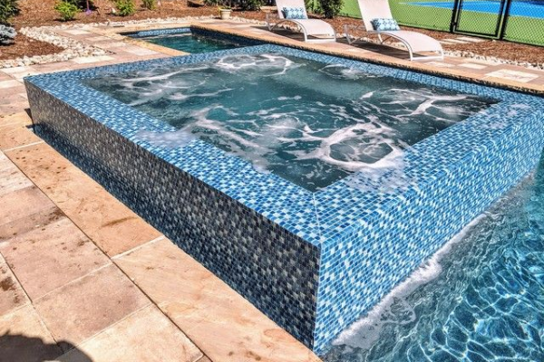 Blue-tiled spa pool with bubbling water beside a larger swimming pool and patio chairs