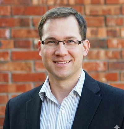 Smiling man in glasses and dark blazer standing before a brick wall