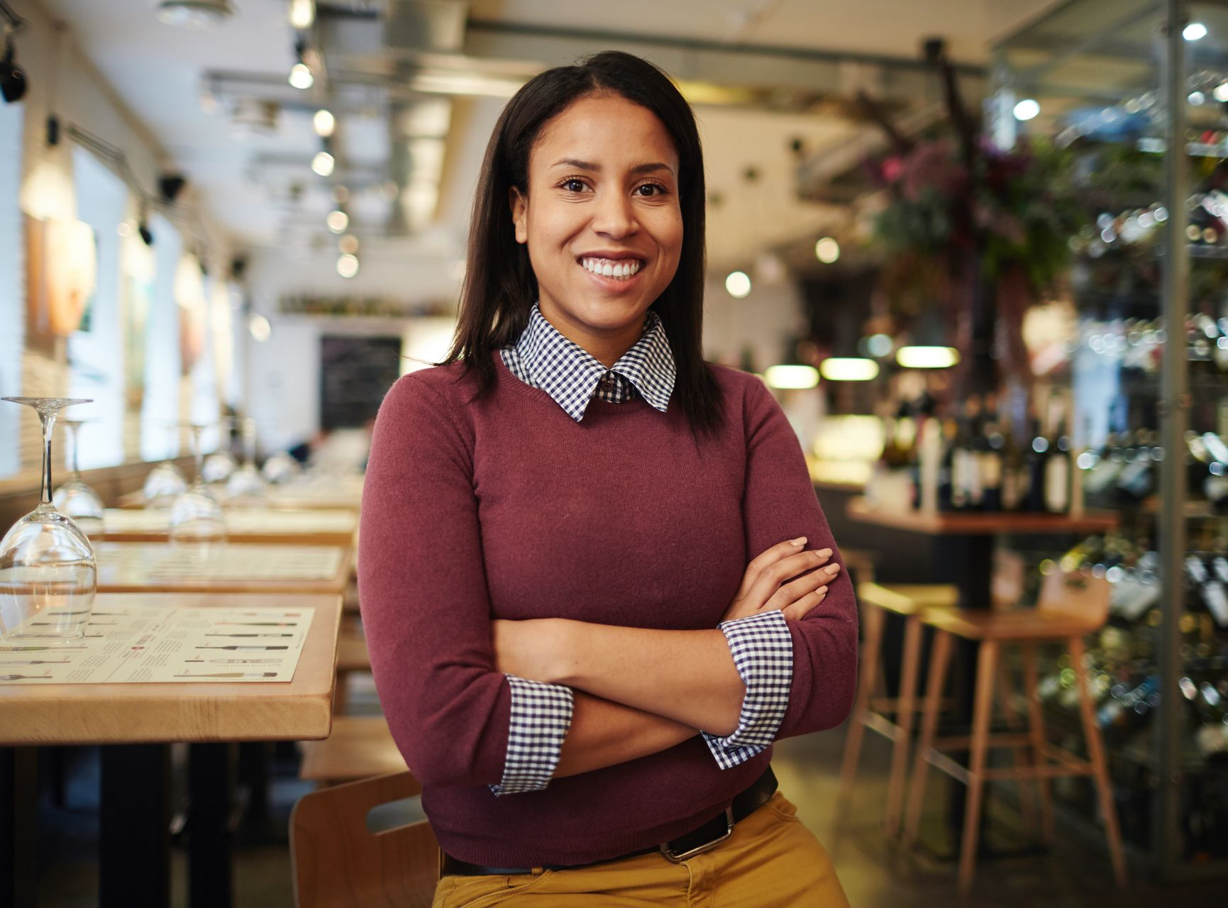 Smiling person in a burgundy sweater stands with arms crossed in a bright café.